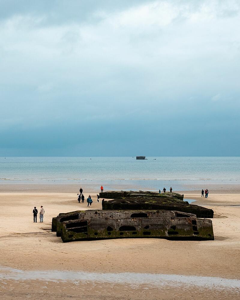 [Translate to Espagnol:] Wide shot of a sandy beach under an overcast sky, where tourists walk around large concrete blocks, an excursion easily accessible from Les Castels campings in Normandy.
