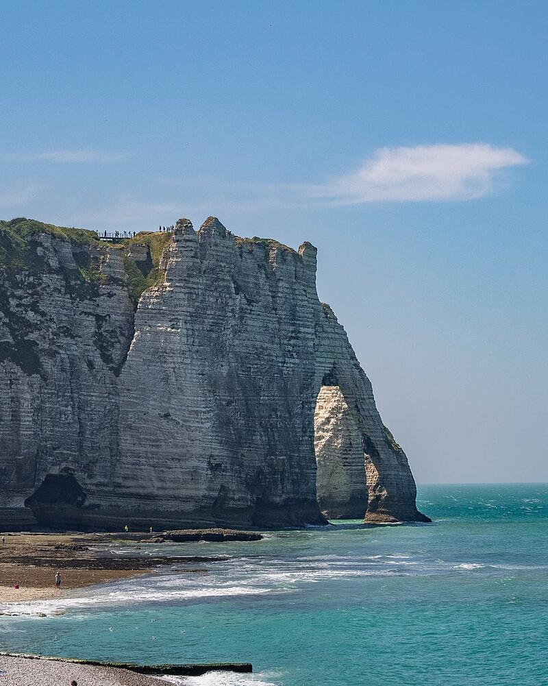 [Translate to Espagnol:] A vertical shot of the famous towering white chalk cliffs and natural arch of Étretat, with waves crashing onto the beach, located near a Les Castels camping resort.