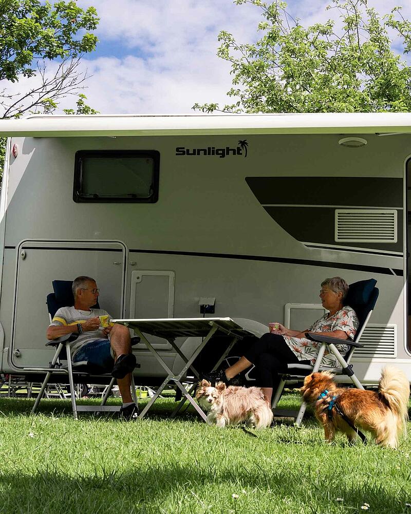 [Translate to Espagnol:] A man and a woman sitting at a camping table under the awning of a modern Sunlight motorhome on a pet-friendly pitch at Les Castels.