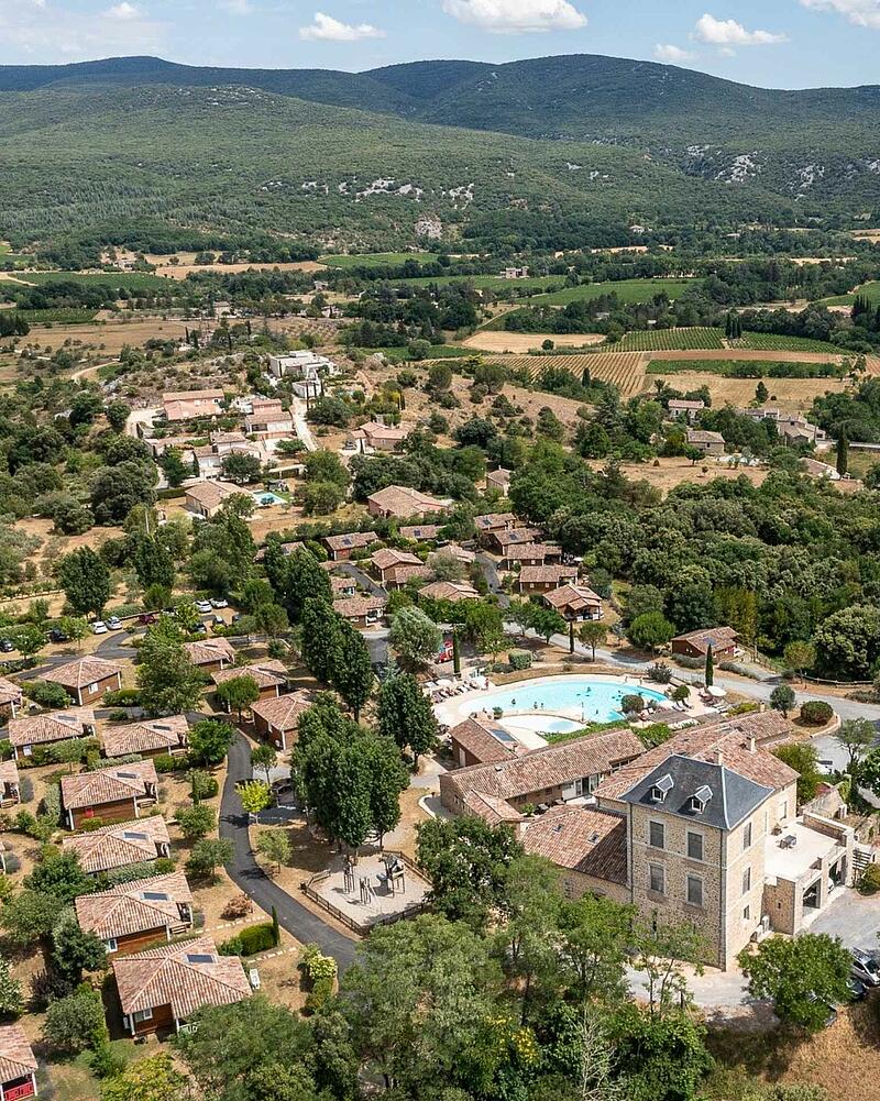 [Translate to Espagnol:] Drone perspective of the heated outdoor swimming pool, stone estate building, and eco-friendly wooden chalets surrounded by nature at a 5-star Les Castels campsite.
