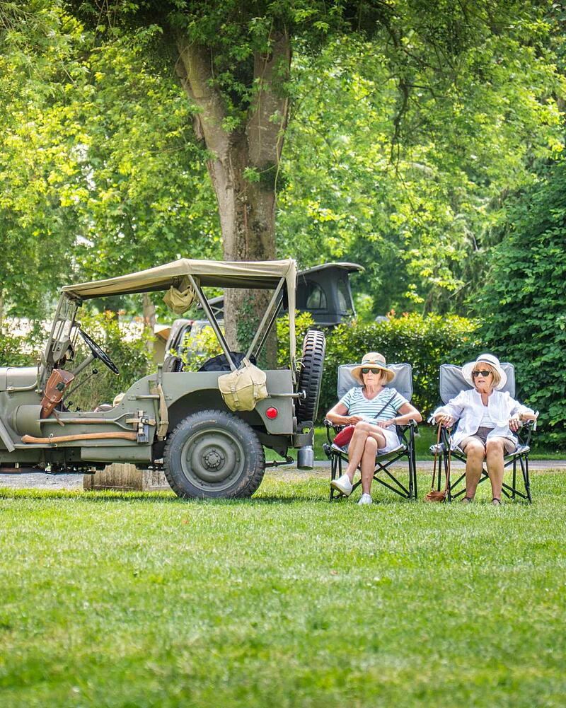 [Translate to Espagnol:] Two senior women wearing sun hats sitting on camping chairs on a green lawn next to a classic green vintage military jeep at a Les Castels campsite.