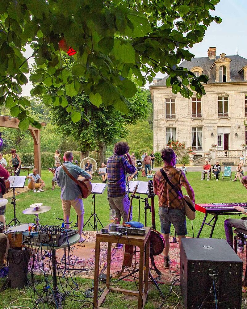 [Translate to Espagnol:] Stage view from behind a live band playing guitars and keyboards outdoors for an audience seated on the grass during a premium evening event at Les Castels.