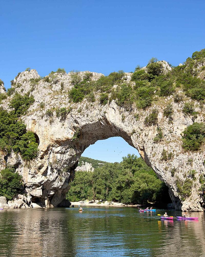[Translate to Espagnol:] Colorful holidaymakers paddling on kayaks and canoes on the river right underneath the giant natural stone bridge of Pont d Arc near Les Castels luxury campsites.