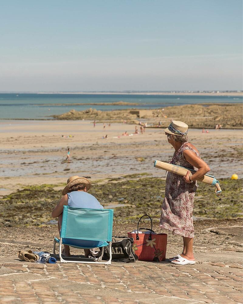 [Translate to Espagnol:] Two women wearing straw hats spending a sunny day on a stone seawall, overlooking a vast sandy beach during a seaside holiday with Les Castels campings.