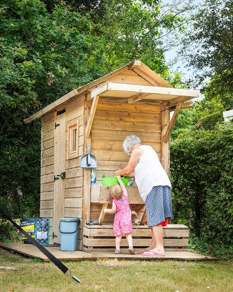 [Translate to Espagnol:] Woman and young girl interact at a rustic wooden outdoor kitchen station on a Les Castels camping estate.