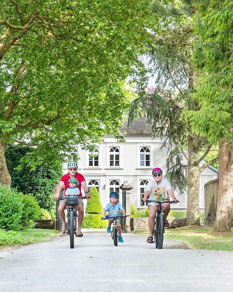 [Translate to Espagnol:] A family of four, including two young children wearing helmets, cycling towards the camera on a gravel path during their holiday at a Les Castels domain.