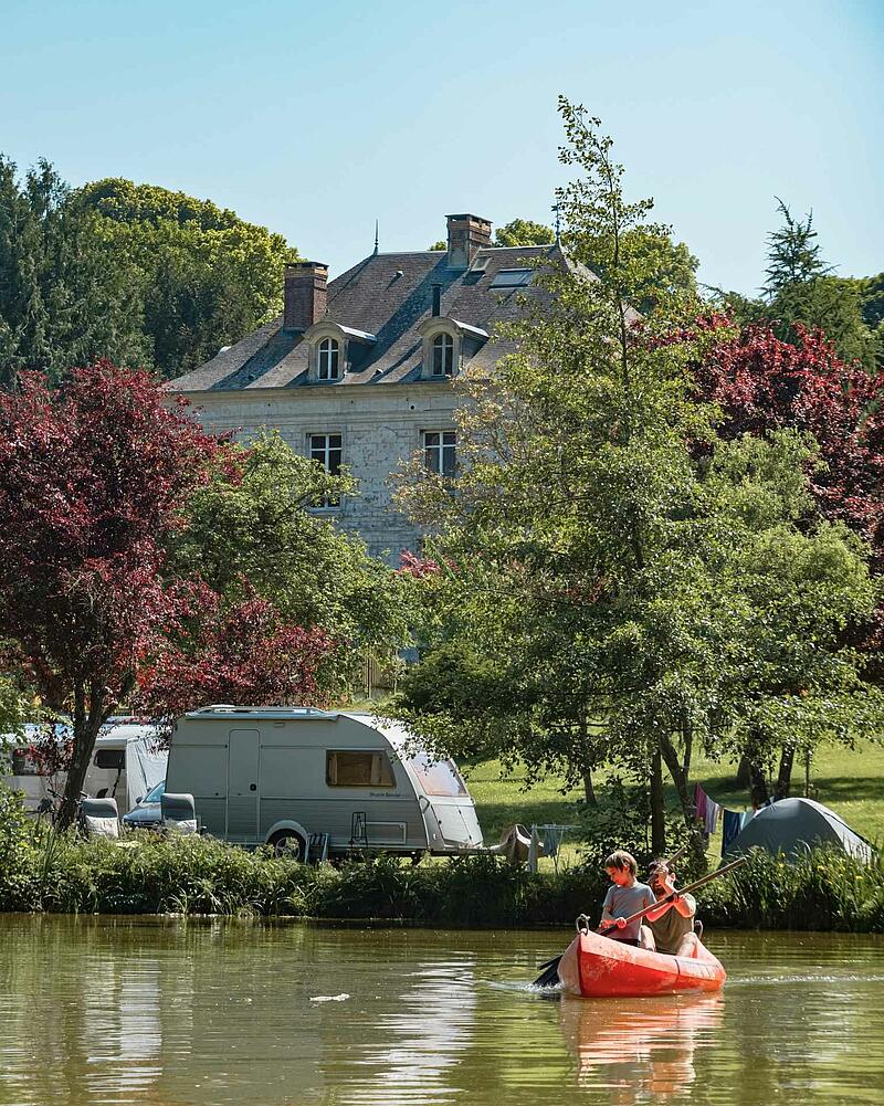 [Translate to Espagnol:] An orange canoe with a man and a young boy paddling on a calm lake, with a caravan parked on the grassy bank of a Les Castels estate.