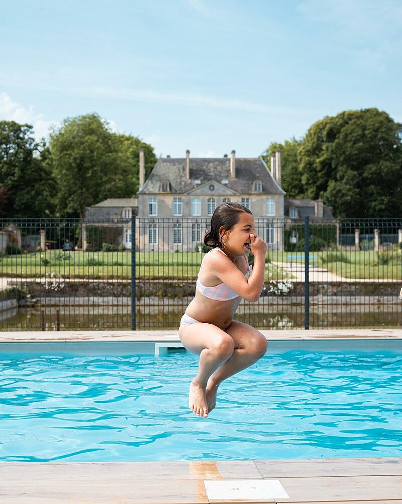 [Translate to Espagnol:] A young girl mid-air in a cannonball jump into a crystal-clear outdoor swimming pool, located in the gardens of a grand French estate at Les Castels.