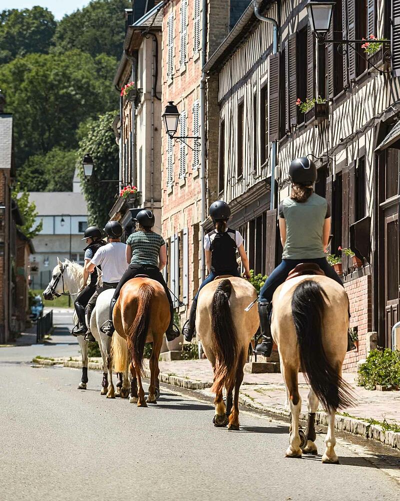 [Translate to Espagnol:] A group of riders on horseback walking down a village street lined with classic half-timbered stone houses, an outdoor activity proposed near your Les Castels camping resort.