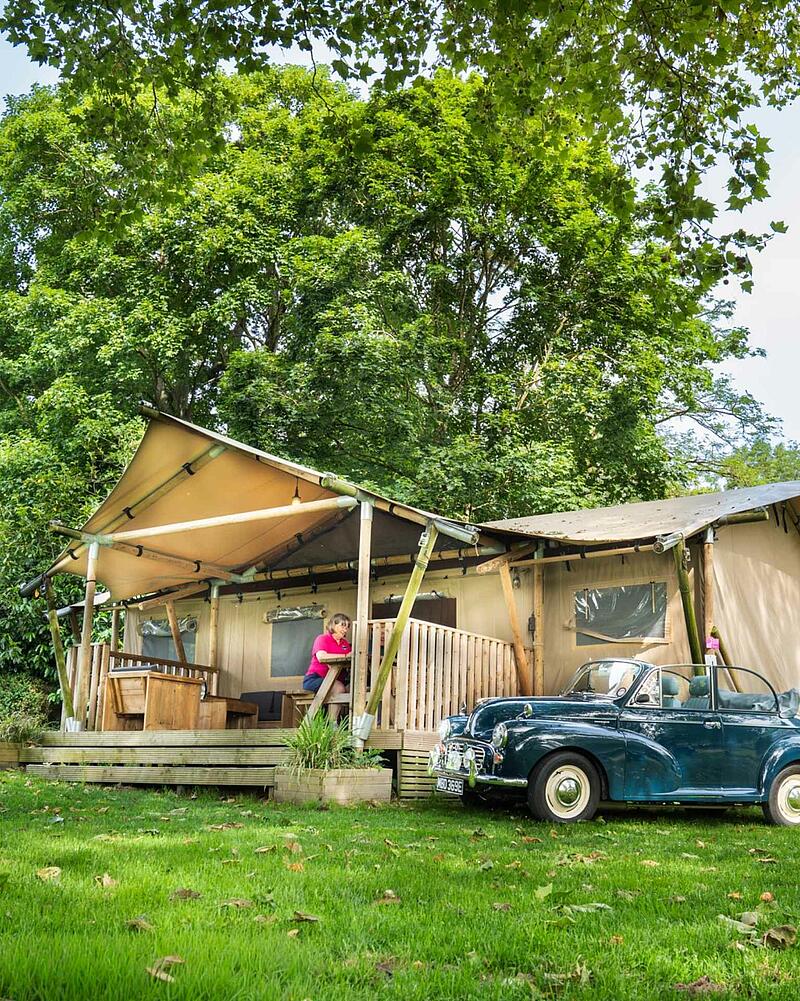 [Translate to Espagnol:] A large canvas glamping safari tent with a wooden porch set amongst lush green trees on a premium pitch at a Les Castels 5-star campsite.