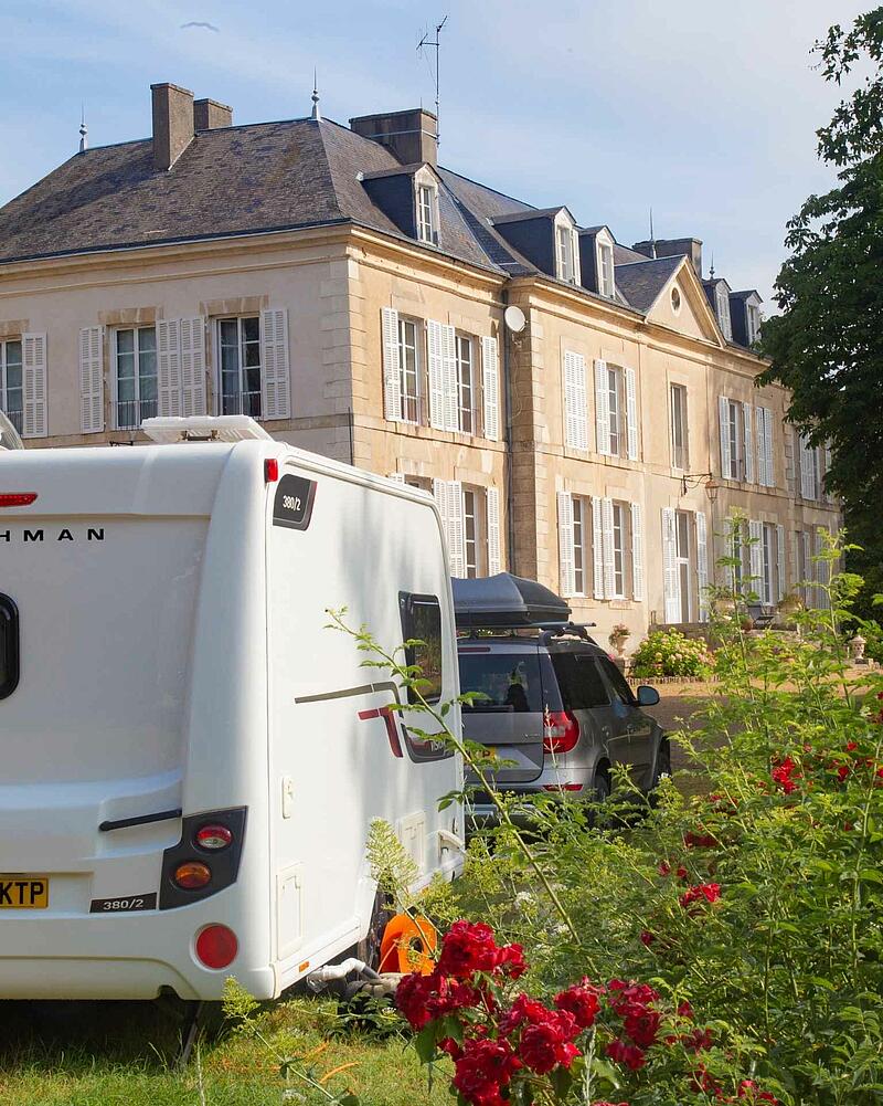 [Translate to Espagnol:] The back of a white caravan and a grey SUV parked on a sunny grass pitch in front of a grand, historic stone chateau, beautifully framed by blooming red roses in the foreground.