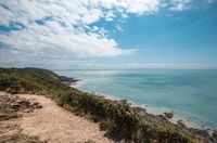 [Translate to Espagnol:] A scenic coastal view in Normandy featuring a dirt pathway on a grassy cliff overlooking a vast expanse of turquoise sea under a blue sky with fluffy white clouds, perfect for an outdoor stay at Les Castels.